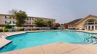 Located at 1025 Kensington Dr in Jacksonville, North Carolina, this outdoor community amenity space offers a well-appointed recreational area designed for residents to relax and socialize. At the heart of the space is a generously sized swimming pool with a curved, freeform shape, featuring clearly marked depth indicators and equipped with stainless steel entry rails for easy access. The surrounding concrete sun deck provides ample room for lounging, and is furnished with multiple blue lounge chairs and umbrellas positioned throughout, offering both sun and shade options. A white perimeter fence encloses the pool area, creating a defined and secure environment. Adjacent to the pool is a clubhouse building with a distinctive gabled roofline and warm yellow siding, providing a dedicated facility for community use. Mature trees and ornamental plantings, including flowering varieties, add natural beauty and shade to the landscape. A playground area is visible beyond the pool enclosure, offering recreational space for younger residents. The multi-story residential buildings, featuring classic brick and neutral-toned exteriors with black-shuttered windows, frame the backdrop of the amenity area, contributing to a cohesive and inviting community atmosphere.