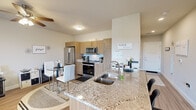 This thoughtfully designed single-floor studio offers a seamless open-concept layout that integrates the kitchen, dining, and living areas into one cohesive space. The kitchen is a standout feature, showcasing warm wood-tone cabinetry, granite countertops with a double-basin sink, a subway tile backsplash, and a full suite of stainless steel appliances including a French door refrigerator, electric range, and over-the-range microwave. A kitchen island provides additional prep space and casual seating, while a compact dining area with a round glass-top table and white chairs sits conveniently nearby. Light wood-tone flooring flows consistently throughout the space, contributing to a clean and contemporary aesthetic. The living area is bright and functional, anchored by a large sliding glass door that fills the room with natural light and opens onto a private balcony, where a glass-panel railing frames an expansive view of the surrounding greenery and landscape. Track lighting and flush-mount ceiling fixtures provide ample illumination throughout. The overall ambiance is modern and efficient, balancing practical design with stylish finishes that make the most of the available space.