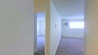 This single-floor space presents a simple, functional layout characterized by clean white walls and wall-to-wall carpeting throughout. The interior features an open corridor that connects multiple rooms, with a doorway leading into an adjoining space that benefits from natural light filtering through a window. The main area is generously proportioned, providing a comfortable sense of openness, while the neutral white walls and light-colored ceiling create a bright, airy atmosphere. A window unit is visible along one wall, supplementing climate control within the space. The carpeted flooring in a muted tone runs consistently through the rooms, contributing to a cohesive and understated aesthetic. Electrical outlets are positioned along the walls, and the overall layout appears straightforward and adaptable for various uses. The space is move-in ready in terms of its structural finishes, offering a blank canvas that can accommodate a range of furniture arrangements and functional needs. The minimal detailing throughout keeps the focus on practicality and usability.