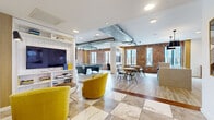 This thoughtfully designed amenity lounge spans a generous single floor, blending industrial aesthetics with contemporary comfort across several distinct functional zones. Exposed brick walls serve as a defining architectural feature throughout, complemented by visible metallic ductwork along the ceiling that reinforces the space's industrial-chic character. Recessed lighting and multiple windows provide consistent natural light, offering views of the surrounding cityscape.

The lounge area near the entrance features bold yellow velvet swivel chairs arranged around a low side table, anchored by a built-in white shelving unit housing a large flat-screen television. A checkered marble-tile floor distinguishes this zone from the rest of the space. Transitioning inward, light wood-look flooring with rich wood inlay accents connects the remaining areas. A billiards table occupies the central open area, while multiple round dining tables with modern black chairs provide casual gathering space.

A tuck-in booth with teal upholstered seating offers an intimate alternative seating nook. The kitchen area is a standout feature, showcasing a substantial quartz waterfall island with black leather bar stools, a dramatic marble-tiled backsplash, warm wood cabinetry, stainless steel appliances, and statement pendant lighting. Together, these elements create a versatile, well-appointed communal space.