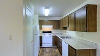 This single-story space includes a well-organized kitchen that serves as a central feature. The kitchen showcases warm, wood-toned cabinetry lining both the upper and lower walls, providing ample storage throughout. Light-colored countertops offer generous workspace, complementing the cabinetry in a classic, practical aesthetic. White appliances—including a freestanding electric range with an overhead range hood, a full-sized refrigerator, and a dishwasher—are neatly integrated into the layout. The kitchen is arranged in a U-shaped configuration, maximizing usable counter and storage space while maintaining an efficient workflow. A stainless steel sink is positioned along one wall, adding a functional focal point. Hardwood-style flooring runs throughout the kitchen, lending warmth and durability underfoot. Overhead, a flush-mounted fluorescent light fixture provides bright, even illumination across the space. The overall ambiance of the kitchen is practical and straightforward, with a classic design sensibility suited to everyday living.