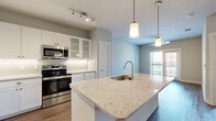 This single-floor residence is designed with a modern, open-concept layout that seamlessly connects the kitchen, dining, and living areas. The kitchen serves as a functional centerpiece, featuring a large granite-topped island with an undermount sink and gold-toned faucet, illuminated by stylish pendant lighting. White shaker-style cabinetry with glass-front upper panels lines the walls, complemented by a white subway tile backsplash, under-cabinet lighting, and a full suite of stainless steel appliances including a refrigerator, range, microwave, and dishwasher. Track lighting above further enhances the kitchen's brightness.

Wood-look plank flooring extends throughout the main living areas, adding warmth and continuity. The living area benefits from natural light and direct access to a private balcony through a glass-panel door, with a ceiling fan overhead. Additional rooms feature plush carpeting and generous windows that admit abundant natural light. The bathroom area includes wood-look flooring, a white vanity, and a bathtub with a window above. Neutral-toned walls throughout the unit create a clean, cohesive aesthetic well-suited for a variety of furnishing styles.
