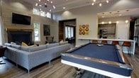 This single-story space is thoughtfully designed for entertaining, combining modern finishes with warm, rustic accents throughout. The main living and game area is anchored by a sleek pool table with a navy felt surface and wood trim, positioned beneath a striking Sputnik-style chandelier that serves as a bold focal point. Elevated ceilings enhance the sense of openness, while large windows allow natural light to fill the space.

Adjacent to the game area, a comfortable lounge features a generous sectional sofa and an accent chair with southwestern-patterned cushions, arranged near a stone fireplace. Colorful vinyl record-inspired wall art adds a playful, eclectic touch to the neutral-toned walls.

A doorway framed by reclaimed wood paneling leads to an additional seating or lounge area, visible in the background, extending the entertaining footprint of the space. The open kitchen is equipped with stainless steel appliances, dark cabinetry, stone backsplash, and a breakfast bar with pendant lighting overhead. Wood-look flooring runs throughout, tying all areas together with a cohesive, contemporary-rustic aesthetic.