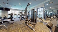This single-floor fitness center offers a thoughtfully designed workout environment packed with professional-grade equipment. The space is outfitted with a wide array of strength-training machines, including Matrix cable systems and various selectorized weight machines, alongside a dedicated free weights area featuring a full dumbbell rack and adjustable benches. The cardio zone is well-represented with treadmills, elliptical trainers, and stationary bikes positioned along the perimeter, benefiting from an abundance of natural light streaming through large, blue-trimmed windows that overlook lush greenery outside. Floor-to-ceiling mirrors line multiple walls, visually expanding the space and allowing users to monitor their form throughout their workouts. The interior is finished with elegant crown molding along the ceiling, recessed lighting, and a patterned carpet in warm tones of gold, gray, and brown that adds a refined touch to the athletic setting. A water station and medicine balls are also available for added convenience. Access to the fitness center is provided through blue-framed glass doors, which connect to adjacent amenity spaces. The overall ambiance is clean, modern, and welcoming, making it a well-rounded facility suitable for a full range of fitness routines.