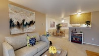 This single-floor residence offers a thoughtfully designed open-concept layout that seamlessly connects the living, dining, and kitchen areas into one cohesive and functional space. The living area is warm and welcoming, anchored by plush carpeted flooring and neutral beige walls that create a calm, inviting ambiance. A large statement artwork adds a bold visual focal point, complementing the comfortable seating arrangement centered around a rustic wood coffee table. The space flows naturally into the dining area, where a ceiling fan with a light fixture provides both comfort and illumination above a wooden dining table and chairs. Colorful artwork adorns the dining area walls, adding personality and vibrancy to the space. Adjacent to the dining area, the kitchen is partially open to the main living space via a raised bar-style counter, allowing for easy interaction between the rooms. Wood-toned cabinetry and under-cabinet lighting give the kitchen a classic, functional character. A charming painted accent cabinet near the kitchen entry adds an eclectic, decorative touch. Throughout the space, the consistent use of soft carpet, warm wall tones, and curated décor creates a cohesive and livable environment that feels both comfortable and stylish.