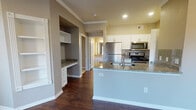 This single-floor residence offers a thoughtfully designed layout with a blend of practical features and modern finishes throughout. The open-concept kitchen is a standout, showcasing granite countertops, white cabinetry, a stainless steel refrigerator, microwave, and range, along with a peninsula that provides additional prep and seating space. A tasteful tile backsplash adds a decorative touch, while recessed lighting ensures the area is well-lit. Adjacent to the kitchen is a built-in desk nook with granite countertop and a set of white open shelving, offering a convenient workspace or display area. The living room is generously sized, featuring rich dark hardwood-style flooring, crown molding, a ceiling fan, and multiple windows dressed with blinds that allow for ample natural light and outdoor views. The entryway is clean and welcoming, with the same dark flooring extending seamlessly throughout the main living areas. The full bathroom is well-equipped with a dual-sink vanity topped with granite, a large mirror, bright vanity lighting, white cabinetry, and a tub-shower combination. The bedroom features plush carpet flooring, a ceiling fan, a window for natural light, and access to a spacious walk-in closet with wire shelving. Neutral wall tones and consistent trim work give the entire space a cohesive, polished appearance.