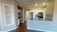 This single-floor residence offers a thoughtfully designed layout with a blend of practical features and modern finishes throughout. The open-concept kitchen is a standout, showcasing granite countertops, white cabinetry, a stainless steel refrigerator, microwave, and range, along with a peninsula that provides additional prep and seating space. A tasteful tile backsplash adds a decorative touch, while recessed lighting ensures the area is well-lit. Adjacent to the kitchen is a built-in desk nook with granite countertop and a set of white open shelving, offering a convenient workspace or display area. The living room is generously sized, featuring rich dark hardwood-style flooring, crown molding, a ceiling fan, and multiple windows dressed with blinds that allow for ample natural light and outdoor views. The entryway is clean and welcoming, with the same dark flooring extending seamlessly throughout the main living areas. The full bathroom is well-equipped with a dual-sink vanity topped with granite, a large mirror, bright vanity lighting, white cabinetry, and a tub-shower combination. The bedroom features plush carpet flooring, a ceiling fan, a window for natural light, and access to a spacious walk-in closet with wire shelving. Neutral wall tones and consistent trim work give the entire space a cohesive, polished appearance.