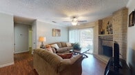 This single-story home offers a thoughtfully arranged layout with multiple living spaces designed for both comfort and functionality. The living room serves as the focal point, featuring rich hardwood-style flooring, a ceiling fan with light fixture, and a striking floor-to-ceiling brick fireplace that anchors the room. Large sliding glass doors with vertical blinds flood the space with natural light and provide access to an outdoor area, offering pleasant exterior views.

Adjacent to the living room, the dining area accommodates a full-sized table and chairs beneath a pendant light fixture, with a window that allows additional natural light. Crown molding adds a refined detail throughout these shared spaces.

The kitchen is equipped with white cabinetry, granite-style countertops, stainless steel appliances, and hardwood-style flooring, with convenient access to an in-unit laundry area.

The home includes two bedrooms, each carpeted and featuring ceiling fans, textured ceilings, and ample closet space. The primary bedroom includes mirrored sliding closet doors and room for a full dresser arrangement. The second bedroom is cozily appointed with warm neutral tones and decorative wall accents. Throughout, the home maintains a consistent, welcoming aesthetic.