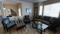 This single-floor space showcases a thoughtfully designed living area that balances contemporary style with bold decorative choices. The living room features rich, dark hardwood flooring that anchors the space and provides a striking contrast against neutral, warm-toned walls. Multiple windows fitted with white blinds allow generous natural light to fill the room, enhancing its open and welcoming atmosphere.

The living area is furnished with a cohesive arrangement that includes a dark-framed sofa, an accent chair with a zebra-print cushion, and a matching ottoman with a geometric patterned upholstery. A glass-top coffee table and a smaller side table with dark metal frames add a modern touch, while a zebra-print area rug defines the seating zone with visual impact. Decorative elements include tall gold vases, dried branches, and a framed full-length mirror, all contributing to the space's eclectic yet curated aesthetic.

An abstract teal and blue wall canvas adds a pop of color against the neutral backdrop. A glimpse into an adjacent area suggests additional rooms beyond, hinting at a functional and connected floor plan throughout the space.