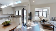 This well-appointed two-story space showcases a thoughtfully designed open-concept layout that effortlessly connects the kitchen, dining, and living areas into a cohesive, functional whole. The interior is characterized by soaring vaulted ceilings that amplify the sense of volume and airiness, while clerestory windows positioned at height allow an abundance of natural light to pour into the space throughout the day.

The kitchen features crisp white cabinetry, clean countertops, and a practical layout that flows naturally into the dining area, where a round wooden table and contemporary white chairs create an inviting setting. Dark-framed windows throughout the space provide a striking modern contrast against the bright white walls, lending an architectural sophistication to the overall aesthetic.

A set of sleek black French doors opens directly to an outdoor area, establishing a seamless connection between the interior and exterior. The living area is finished with rich dark hardwood flooring that grounds the neutral palette, complemented by a comfortable seating arrangement and a media console. Natural light streams in from multiple directions, casting warmth across the open floor plan. The combination of high ceilings, contemporary finishes, and an abundance of natural light creates a bright, modern, and welcoming atmosphere throughout the entire space.