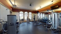 This dedicated fitness center is designed with both functionality and aesthetics in mind, presenting a sleek black-and-white color scheme that creates a sharp, professional atmosphere. The space is anchored by a robust selection of cardio equipment, including multiple treadmills, elliptical trainers, stair climbers, and upright and recumbent stationary bikes — many equipped with individual monitor screens for an enhanced workout experience. Large windows line the perimeter walls, fitted with blinds that allow ample natural light to fill the room while maintaining privacy. Ceiling fans complement the recessed lighting overhead, ensuring a comfortable environment during intense workouts. Full-length mirrors extend along one wall, visually expanding the space and allowing users to monitor their form. The flooring consists of durable dark rubber gym matting, providing both safety and sound absorption throughout. A storage area along one wall offers cubbies and shelving for personal belongings, while a weight and exercise accessory section — including stability balls and weight plates — supports a variety of training routines. Glass-paneled doors lead directly to an outdoor courtyard area with lush greenery, offering a refreshing connection to the outdoors.