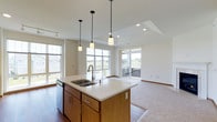 This single-floor residence offers an open-concept layout that seamlessly connects the kitchen and living areas. The kitchen serves as the heart of the space, anchored by a central island with a white countertop, double stainless steel sink, and warm wood cabinetry. Three pendant lights hang above the island, providing focused task lighting that complements the recessed ceiling lights throughout. A dishwasher is integrated beneath the island, adding functional convenience.

The kitchen flows naturally into the living area, where durable hardwood flooring transitions to plush carpeting. A tray ceiling detail adds architectural interest and a sense of height overhead. Multiple large windows line the walls, filling the space with abundant natural light and offering open views of the surrounding landscape.

A sliding glass door leads to a private balcony, which overlooks a scenic streetscape bordered by lush greenery, open fields, and mature trees. The balcony extends the living experience outdoors, providing a peaceful setting.

Overall, the space presents a clean, neutral palette with modern finishes, creating a bright and welcoming atmosphere well-suited for comfortable, everyday living.