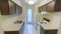 This single-floor residence offers a practical and well-organized layout designed for comfortable everyday living. The heart of the home is a classic galley-style kitchen, efficiently arranged with cabinetry and counter space running along both sides, maximizing storage and functionality. Dark wood upper and lower cabinets provide generous storage capacity, while the light-toned countertops offer ample workspace for meal preparation. The kitchen is equipped with a full suite of white appliances, including a coil-burner electric range with a ventilation hood, a full-size refrigerator, and a sink with a single-basin setup. Light tile flooring runs the length of the kitchen, contributing to a clean and easy-to-maintain environment. A standout feature of the kitchen is the door at the far end, fitted with a window that invites natural light deep into the space and offers a view of the outdoors, creating an airy and open feel despite the galley configuration. Overhead, a ceiling-mounted light fixture provides consistent illumination throughout. The neutral, light-colored walls keep the space feeling bright and welcoming, providing a versatile backdrop suited to a variety of interior styles.