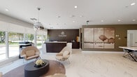 Located at 9609–9689 SW 152nd St in Miami, FL, the AHS Coral Reef Rental Community leasing office is a single-story space designed with a polished, contemporary aesthetic that reflects the caliber of the community it represents. The interior is anchored by expansive marble-look porcelain flooring that runs throughout, lending a refined and cohesive feel to the entire space.

Upon entry, visitors are welcomed by a dedicated reception area featuring a dark wood desk prominently displaying the AHS Coral Reef branding. A large-scale sepia-toned artwork depicting a historic airship scene serves as a striking decorative focal point nearby. The reception zone flows seamlessly into a comfortable lounge area furnished with sculptural upholstered chairs, a round coffee table, and a cowhide-style area rug, all illuminated by a distinctive spherical chrome pendant light.

Floor-to-ceiling windows along the perimeter allow generous natural light to fill the space while offering views of the surrounding exterior. Decorative topiary arrangements add organic texture near the entry points. A glass-enclosed conference room provides a semi-private meeting area equipped with a wall-mounted display screen and seating for several guests, maintaining visual openness while offering functional separation. The overall ambiance is professional, bright, and welcoming.