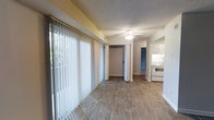 This single-story space showcases an open-concept layout that seamlessly connects the main living area to the kitchen. The living area is finished with wood-style laminate flooring that runs throughout, lending warmth and continuity to the space. A ceiling fan is centrally positioned overhead, providing both air circulation and ambient lighting. The textured ceiling adds a classic residential character to the interior.

A prominent wall of vertical blinds lines one side of the living area, covering large windows that invite natural light deep into the space. The greenery visible through the windows hints at a pleasant outdoor setting.

An open doorway leads to an adjacent room, offering flexible use of the layout. The kitchen, visible from the main living area, features white cabinetry, a white electric range, and a clean, functional design. White walls and trim throughout the space create a bright, neutral palette that complements the wood-tone flooring.

Overall, the space presents a clean, practical layout with a bright and open ambiance, well-suited for everyday living.