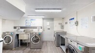Located at 1660 Bathurst Street in Toronto, this shared laundry room is a well-equipped, single-floor facility designed to serve building residents efficiently. The room features a clean, utilitarian aesthetic with white-painted walls — some finished in brick — and durable wood-look flooring throughout that gives the space a tidy, maintained appearance.

The laundry area is outfitted with multiple front-loading washers and dryers in a modern stainless steel finish, arranged along the walls to maximize usability. Generous stainless steel folding tables are positioned alongside the machines, providing ample workspace for sorting and folding laundry. A deep utility sink is also available for hand-washing or pre-treating garments.

A digital payment kiosk is installed, offering a coin-free payment option for added convenience. Instructional signage for washer and dryer usage, as well as dryer safety guidelines, is clearly posted throughout the room. Overhead fluorescent lighting ensures the space is bright and well-lit.

Safety is addressed with a mounted fire extinguisher near the entrance and a clearly marked emergency exit door. A security camera is also present, contributing to a safe environment. The room is accessed through a dedicated door with an exit sign, keeping it organized and separated from other building areas.