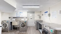 Located at 1660 Bathurst Street in Toronto, this shared laundry room is a well-equipped, single-floor facility designed to serve building residents efficiently. The room features a clean, utilitarian aesthetic with white-painted walls — some finished in brick — and durable wood-look flooring throughout that gives the space a tidy, maintained appearance.

The laundry area is outfitted with multiple front-loading washers and dryers in a modern stainless steel finish, arranged along the walls to maximize usability. Generous stainless steel folding tables are positioned alongside the machines, providing ample workspace for sorting and folding laundry. A deep utility sink is also available for hand-washing or pre-treating garments.

A digital payment kiosk is installed, offering a coin-free payment option for added convenience. Instructional signage for washer and dryer usage, as well as dryer safety guidelines, is clearly posted throughout the room. Overhead fluorescent lighting ensures the space is bright and well-lit.

Safety is addressed with a mounted fire extinguisher near the entrance and a clearly marked emergency exit door. A security camera is also present, contributing to a safe environment. The room is accessed through a dedicated door with an exit sign, keeping it organized and separated from other building areas.