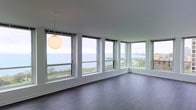 This single-floor space offers a striking blend of modern design and breathtaking natural scenery. The main living area is defined by a wraparound bank of large, white-framed windows that flood the interior with abundant natural light and frame unobstructed panoramic views of a vast body of water and the surrounding cityscape beyond. The elevated vantage point enhances the sense of openness, making the horizon a constant visual centerpiece from virtually every angle of the room.

The interior features rich, dark hardwood flooring that runs throughout, providing a warm contrast to the crisp white walls and window trim. The clean lines and neutral palette create a minimalist, contemporary atmosphere that feels both sophisticated and inviting. A single spherical pendant light hangs from the smooth, white ceiling, adding a subtle decorative focal point without detracting from the architectural simplicity of the space.

The open layout allows for flexible use and arrangement, maximizing the impact of the surrounding views. The corner window configuration is a standout architectural element, drawing the eye outward and reinforcing the connection between the interior and the expansive outdoor scenery. Overall, this space delivers a serene, light-filled environment with an exceptional waterfront outlook.