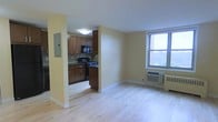 This single-floor unit features an open layout that connects the living area and kitchen in a seamless, functional design. The light hardwood flooring extends throughout, lending warmth and continuity to the space. Pale yellow walls contribute to an airy, welcoming atmosphere, while a single window in the living area admits natural light and offers an exterior view.

The kitchen is tucked into a semi-enclosed alcove defined by a structural column, providing a sense of separation while maintaining openness to the main living space. It is equipped with rich dark wood cabinetry, dark countertops, a black gas range, and a built-in microwave above the stove, offering a complete cooking setup within a compact footprint. A utility panel is mounted on the adjacent wall near the kitchen entrance.

The living area is generously proportioned and unobstructed, allowing flexible furniture arrangements. A wall-mounted HVAC unit is positioned beneath the window, ensuring year-round climate control. The overall design balances practicality with a clean, understated aesthetic throughout.