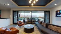 Located along SE Grand Avenue in Portland's vibrant inner eastside, this thoughtfully designed amenity suite offers a polished blend of collaborative and private spaces. The open main area features polished concrete floors, a dramatic black coffered ceiling with recessed lighting, and a large communal dining table surrounded by warm cognac leather chairs. A full kitchenette anchors one wall, outfitted with wood cabinetry, a tile backsplash, stainless steel appliances, and ample counter space, making it well-suited for catered events or everyday use. Adjacent to the kitchen, a media lounge with a mounted display and bold bokeh-inspired wall mural adds a dynamic visual element to the space. The lounge area features a tray ceiling with statement globe pendants, plush seating including a sectional sofa and leather accent chairs, and floor-to-ceiling glass walls that open directly onto a furnished outdoor terrace with urban views. Two private conference rooms, each appointed with solid wood tables, upholstered seating, dark wood-slat ceilings, and integrated lighting, provide quiet settings for focused meetings. Together, these spaces offer a refined and functional environment ideal for residents or tenants seeking premium shared amenities.