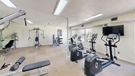 Located at 100 Belle Fontaine Dr in Lafayette, Louisiana, this fitness center offers a well-appointed, single-floor exercise room designed to support a comprehensive workout routine. The space is generously sized, providing ample room for both cardio and strength training activities without feeling cramped.

The exercise room features a variety of cardio equipment, including multiple treadmills, elliptical trainers, and stationary bikes, catering to a range of aerobic workout preferences. Complementing the cardio section, the room is equipped with several strength training machines, including multi-function cable systems and resistance stations, as well as free weights for targeted muscle conditioning.

Natural light fills the space through large windows and glass French doors that open to an outdoor view, creating a bright and energizing workout environment. The textured ceiling with recessed fluorescent lighting ensures consistent illumination throughout the room. Light-toned carpet covers the floor, contributing to a clean and neutral aesthetic, while the soft green and cream-colored walls add a calm, welcoming ambiance.

The layout is open and functional, with clear pathways between equipment stations. A wall-mounted television is also present, adding convenience for users during their workout sessions. Overall, this fitness center provides a practical and comfortable setting for a full range of exercise activities.