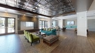 Located at 1000 Mill Road in Ronkonkoma, NY, the Alston Station Square clubhouse is a thoughtfully designed, single-floor amenity space that combines contemporary style with functional versatility. The centerpiece of the space is a grand lounge area anchored by an ornate stamped-metal ceiling with a dark, industrial finish — a striking architectural feature that draws the eye upward and lends the room a distinctive character. Rich wood-look tile flooring flows throughout, unifying the various zones seamlessly.

The main lounge accommodates multiple seating arrangements, including vibrant teal and green upholstered sofas and chairs, tufted ottomans, and accent tables, all oriented around a large flat-screen display. A billiards table occupies one wing, complemented by a bar counter with mosaic tile backsplash and pendant lighting, creating an ideal social hub. Floor-to-ceiling steel-framed glass partition walls define a private lounge nook and allow natural light to pass freely through the space.

Additional areas include a formal dining table with pendant lighting, a curved sectional seating nook, and cozy conversational groupings. Large French doors and oversized windows along the perimeter flood the interior with natural light while offering views of the surrounding outdoor grounds, enhancing the open, welcoming ambiance throughout.