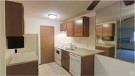 This single-floor residence offers a straightforward, functional layout well-suited for comfortable everyday living. The kitchen is a standout feature, showcasing warm medium-toned wood cabinetry both above and below the countertops, providing ample storage space. White appliances — including an electric range, dishwasher, and refrigerator — complement the neutral countertops and create a clean, cohesive look. The sink is centrally positioned beneath the upper cabinets, making meal prep and cleanup convenient. Light tile flooring runs throughout the kitchen, adding a bright, easy-to-maintain surface underfoot. A ceiling fan is positioned overhead, contributing to air circulation and comfort in the space. A mirrored wall accent near the kitchen adds a sense of depth and openness to the area. A door off the kitchen provides additional access, enhancing the flow and practicality of the floor plan. The overall ambiance of the space is practical and welcoming, with a neutral color palette that offers a versatile backdrop for personalization. The single-floor layout ensures easy accessibility throughout the entire residence.