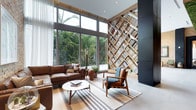 This thoughtfully designed single-floor space makes an immediate impression with its soaring double-height ceilings and an abundance of natural light. The living area is anchored by a stunning feature wall of diagonal, lattice-style built-in bookshelves crafted from warm natural wood, extending from floor to ceiling and serving as both a functional library and a striking architectural statement. Adjacent to the shelving, a deep navy accent panel adds a bold contrast that grounds the composition. Floor-to-ceiling windows and glass sliding doors line the opposite wall, flooding the interior with daylight and framing a lush, verdant garden beyond. The seamless connection between indoors and outdoors creates a calm, open ambiance throughout. Light hardwood flooring runs underfoot, complementing the natural tones of the space, while a striped area rug defines the seating zone. The living area features a rich tan leather sofa and mid-century-inspired wooden armchairs arranged around a round white coffee table — a blend of classic and contemporary furnishings. Sheer curtains soften the light and add an elegant, airy quality. The overall aesthetic is refined yet welcoming, balancing architectural drama with comfortable, livable design.