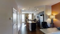 This well-appointed two-story residence offers a thoughtfully designed layout that balances functionality with modern aesthetics. The open-concept main level centers around a striking kitchen, where dark espresso cabinetry provides a bold contrast against light granite countertops and a suite of stainless steel appliances, including a full-size refrigerator, built-in microwave, and range. A central kitchen island with an integrated sink and pendant lighting above serves as both a prep space and a casual dining area, accommodated by bar-style seating. Rich hardwood flooring flows seamlessly throughout the main living area, adding warmth and continuity to the space. Recessed lighting and flush-mount ceiling fixtures ensure the interior is well-illuminated throughout the day. The living area, visible from the kitchen, benefits from natural light streaming through large windows dressed with elegant curtains, creating a bright and welcoming atmosphere. Multiple white paneled doors along the hallway suggest ample storage and closet space. The neutral wall tones and clean architectural lines throughout lend the home a contemporary feel, while the two-floor layout provides additional living or sleeping quarters above, offering a sense of spaciousness and versatility suited to a variety of lifestyles.