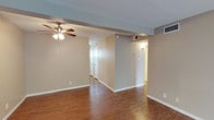 This single-story space presents a straightforward and functional layout characterized by warm hardwood-style flooring that runs throughout the open main area. The neutral beige walls provide a versatile backdrop, complementing the clean white baseboards and trim that frame each room. A ceiling fan with an integrated light fixture is centrally positioned, contributing both airflow and ambient lighting to the main living area.

Structural columns and partition walls divide the space into distinct zones while maintaining a sense of openness, with hallways branching toward additional rooms visible in the background. HVAC vents are integrated into both the walls and ceiling, indicating a comprehensive climate control system throughout. The textured ceiling adds subtle visual depth to the overhead plane.

The hallways lead to what appear to be closeted or private rooms, with white doors providing clean contrast against the neutral walls. Wall-mounted lighting fixtures accent the corridors, providing functional illumination. Overall, the space conveys a clean, move-in ready condition with consistent finishes and a practical, open feel suited for a variety of uses.