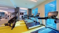 This contemporary fitness center is thoughtfully designed to accommodate a full range of workout needs across a single, elongated floor plan. The space is divided into distinct functional zones, each outfitted with professional-grade equipment. The cardio area features a row of Life Fitness treadmills and stationary bikes positioned along large windows, offering views of the surrounding cityscape and greenery while flooding the floor with natural light. A vibrant yellow flooring accent defines this zone, adding a bold visual contrast to the neutral gray and white palette throughout. Adjacent to the cardio section, a striking yellow multi-functional cable and strength rig serves as the centerpiece of the free weights and functional training area, complemented by stability balls, a punching bag, and open floor space finished with teal exercise mats. The elliptical and stair-climber zone is anchored by a wall-mounted flat-screen television, making workouts more engaging. A dedicated strength and stretching area features striped carpet flooring, floor-to-ceiling mirrors that expand the sense of space, and select selectorized machines. Recessed and linear LED lighting throughout provides consistent, even illumination, while glass partitions and clean architectural lines contribute to the facility's modern, professional ambiance.