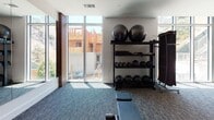 Located at 6711 Sepulveda Blvd in Los Angeles, this well-equipped fitness center is designed to support a full range of workout routines in a single, open-plan floor. The space features a thoughtful layout with distinct zones for cardio training, strength equipment, and functional fitness. Floor-to-ceiling windows along one side of the gym flood the interior with natural light and offer views of the surrounding outdoor environment, creating an energizing atmosphere for workouts.

The cardio zone is stocked with a robust selection of equipment, including treadmills — among them curved, non-motorized models — ellipticals, and Echelon spin bikes. Nearby, a dedicated strength and cable machine section provides multi-functional weight stations and a well-organized free weights area. A functional training nook features a rack stocked with medicine balls, stability balls, and additional accessories, accompanied by a workout bench positioned beneath large windows.

The interior design balances modern aesthetics with practicality, incorporating warm wood-paneled wall accents, recessed lighting, and textured carpet flooring throughout. Structural columns punctuate the open layout while maintaining a spacious feel. A wall-mounted television is integrated into the wood feature wall, adding an entertainment option for users. The overall ambiance is clean, professional, and welcoming, making this fitness center a comprehensive facility for daily training needs.