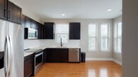 Located at 5010 W Broad St NE in Sugar Hill, Georgia, this single-floor space offers a clean, contemporary design with thoughtful finishes throughout. The open-concept layout is anchored by warm hardwood-style flooring that flows seamlessly through the main living areas, complemented by neutral taupe walls and bright white trim.

The kitchen is a standout feature, showcasing rich dark espresso cabinetry paired with crisp white countertops and a full suite of stainless steel appliances, including a refrigerator with ice maker, electric range, over-the-range microwave, and dishwasher. Multiple windows flood the kitchen with natural light and offer pleasant outdoor views.

The spacious living area continues the hardwood-style flooring and benefits from generous windows fitted with blinds, allowing for flexible light control. The bedroom is finished with plush wall-to-wall carpeting, a ceiling fan, and large windows that invite natural light. A sizable walk-in closet with built-in shelving and hanging rods provides ample storage.

The bathroom is appointed with a glass-enclosed walk-in shower featuring white tile surround and matte black hardware, adding a refined, modern touch. A hallway utility panel and recessed lighting throughout round out the practical and polished character of this well-designed space.