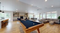The Acero Vancouver clubhouse at 13420 NE Leroy Haagen Memorial Dr is a thoughtfully designed single-story community amenity space offering a range of gathering and recreational areas. Throughout the clubhouse, warm-toned hardwood-style flooring—laid in a striking herringbone pattern in the entry zone—ties the open layout together beneath vaulted and recessed-lit ceilings that lend the space an airy, expansive feel.

The central social area is anchored by a solid wood-framed billiards table and opens into a comfortable lounge featuring a large sectional sofa, a wall-mounted TV, and framed local artwork celebrating Vancouver. French doors provide direct views and access to an outdoor pool area, flooding the interior with natural light. A brick-surround gas fireplace with open rustic shelving adds warmth to the adjacent seating zone, which also includes a dining table and chairs.

The kitchen area is well-appointed with dark cabinetry, a full-length quartz countertop island with bar seating, pendant lighting, a tile backsplash, and appliances including a coffee station. A separate private dining room features a large dark-wood table beneath a geometric pendant light, surrounded by windows overlooking the outdoor amenity space. A welcoming entry nook with patterned accent chairs leads to a glass-door fitness center. The overall atmosphere is contemporary, bright, and community-focused.