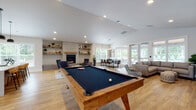 The Acero Vancouver clubhouse at 13420 NE Leroy Haagen Memorial Dr is a thoughtfully designed single-story community amenity space offering a range of gathering and recreational areas. Throughout the clubhouse, warm-toned hardwood-style flooring—laid in a striking herringbone pattern in the entry zone—ties the open layout together beneath vaulted and recessed-lit ceilings that lend the space an airy, expansive feel.

The central social area is anchored by a solid wood-framed billiards table and opens into a comfortable lounge featuring a large sectional sofa, a wall-mounted TV, and framed local artwork celebrating Vancouver. French doors provide direct views and access to an outdoor pool area, flooding the interior with natural light. A brick-surround gas fireplace with open rustic shelving adds warmth to the adjacent seating zone, which also includes a dining table and chairs.

The kitchen area is well-appointed with dark cabinetry, a full-length quartz countertop island with bar seating, pendant lighting, a tile backsplash, and appliances including a coffee station. A separate private dining room features a large dark-wood table beneath a geometric pendant light, surrounded by windows overlooking the outdoor amenity space. A welcoming entry nook with patterned accent chairs leads to a glass-door fitness center. The overall atmosphere is contemporary, bright, and community-focused.