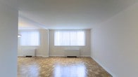 This single-floor space presents a clean and functional layout centered around a generously sized main living area. The room is finished with classic parquet hardwood flooring in a warm honey tone, laid in a traditional block pattern that adds visual texture and character underfoot. The floors appear well-maintained and carry a polished sheen that reflects the natural light streaming in from the large windows along the exterior wall. The windows are fitted with vertical blinds, allowing for adjustable light control while providing a bright, airy atmosphere even when closed. White-painted walls and a smooth white ceiling contribute to the overall sense of openness, keeping the space feeling fresh and neutral. A radiator unit is positioned beneath the main window grouping, indicating a radiant heating system. The minimalist, uncluttered nature of the space makes it highly adaptable for a variety of furniture arrangements and uses. Crown molding details along the ceiling add a subtle architectural refinement to the room. Overall, the space offers a bright, well-lit environment with classic flooring and simple, timeless finishes throughout.