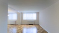 This single-floor space presents a clean and functional layout centered around a generously sized main living area. The room is finished with classic parquet hardwood flooring in a warm honey tone, laid in a traditional block pattern that adds visual texture and character underfoot. The floors appear well-maintained and carry a polished sheen that reflects the natural light streaming in from the large windows along the exterior wall. The windows are fitted with vertical blinds, allowing for adjustable light control while providing a bright, airy atmosphere even when closed. White-painted walls and a smooth white ceiling contribute to the overall sense of openness, keeping the space feeling fresh and neutral. A radiator unit is positioned beneath the main window grouping, indicating a radiant heating system. The minimalist, uncluttered nature of the space makes it highly adaptable for a variety of furniture arrangements and uses. Crown molding details along the ceiling add a subtle architectural refinement to the room. Overall, the space offers a bright, well-lit environment with classic flooring and simple, timeless finishes throughout.