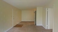 This single-floor space presents a straightforward, functional layout centered around an open living area. The main room features wall-to-wall carpet in a neutral beige tone, creating a clean and understated foundation. Near the front entry door, a tiled transition area provides a practical landing zone, distinguishing the entryway from the carpeted living space. The walls are painted in a soft, warm yellow-beige hue, contributing to an overall light and neutral palette throughout. A textured popcorn ceiling adds a classic finish overhead. The layout includes at least one interior door visible along the far wall, suggesting access to an additional room or hallway beyond the main living space. Electrical outlets are distributed at standard intervals along the base of the walls, and the clean white baseboards provide a crisp contrast to the carpeted flooring. The space is vacant, allowing the architectural simplicity of the layout to be clearly observed. Overall, the space offers a modest, move-in ready environment with a neutral design scheme suited for easy personalization.