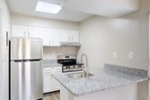 This efficiently designed one-bedroom, one-bathroom unit offers a thoughtful layout across a single floor, making smart use of every square foot. The kitchen serves as a standout feature, boasting granite countertops, crisp white cabinetry, stainless steel appliances including a refrigerator and range, and a peninsula with an integrated sink. A skylight above the kitchen area floods the space with natural light, while a nearby window provides additional brightness and outdoor views. The flooring in the kitchen and entry areas is finished with light wood-tone vinyl planks, lending a clean and contemporary feel. The bedroom is a comfortably sized, carpeted retreat with neutral beige walls and a window dressed with blinds that allow for natural light while maintaining privacy. The room also includes a dedicated closet for storage. The bathroom features tile flooring with a wood-look finish, a full-sized soaking tub with a tiled tub surround, and a curved shower rod. The overall aesthetic throughout the unit is neutral and bright, with soft warm tones on the walls and clean white trim, creating a simple, move-in-ready atmosphere well suited for comfortable everyday living.