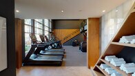 This well-appointed, two-level fitness center offers a comprehensive workout environment designed with both functionality and aesthetics in mind. The space is anchored by a striking design palette of dark charcoal walls, warm wood accents, and neutral flooring, creating a sleek and motivating atmosphere throughout.

The lower level serves as the primary workout floor, housing an extensive row of treadmills and elliptical machines positioned along a dramatic wall of floor-to-ceiling windows. These expansive glazed panels flood the space with natural light and provide open views of the surrounding courtyard. Adjacent to the cardio zone, a dedicated free weights area features a full rack of dumbbells, adjustable benches, and a variety of Hoist-brand strength and resistance machines. Rubber flooring throughout ensures durability and safety during exercise.

A staircase with glass railings connects the lower level to the upper mezzanine floor, which overlooks the main workout area and offers additional equipment and viewing screens. The entry area features stone tile flooring, informational signage, and neatly stocked shelving with fresh towels, providing a polished, amenity-rich welcome. Recessed and linear LED lighting supplements the abundant natural light, ensuring the entire space remains bright and well-illuminated at all hours.