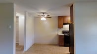 This single-floor residence offers a practical and functional layout with an open connection between the living area and kitchen. The living space features neutral-toned walls, light carpeted flooring, and a ceiling fan with integrated lighting, contributing to a comfortable and well-ventilated atmosphere. The textured ceiling adds a subtle traditional character throughout.

The kitchen is positioned adjacent to the living area and is outfitted with warm-toned wooden cabinetry that provides ample storage. Black appliances, including an electric range and a full-size refrigerator, lend a modern contrast to the lighter countertops. A tile backsplash adds a clean, finished look to the cooking area, while the light-toned flooring offers easy maintenance underfoot.

The partial wall separating the kitchen from the living space defines each area while maintaining an open, connected feel. The overall design is straightforward and well-organized, making efficient use of the available space. The neutral color palette throughout creates a cohesive and inviting environment suitable for everyday living.