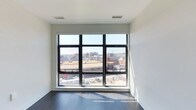 This single-floor space showcases a sleek, contemporary design characterized by clean lines and a minimalist aesthetic. The room features striking floor-to-ceiling windows with black metal frames, arranged in a wide three-panel configuration that floods the interior with natural light and frames an expansive urban cityscape view. The elevated vantage point offers sightlines across rooftops and city buildings, creating a dynamic connection to the surrounding metropolitan environment. Smooth white walls contrast elegantly with rich, dark hardwood flooring, lending the space a sophisticated and modern character. Ceiling-mounted ventilation registers are seamlessly integrated into the white ceiling, maintaining the uncluttered visual aesthetic. The open layout and generous window placement ensure the room feels spacious and airy throughout the day, with sunlight casting natural patterns across the dark floors. The overall ambiance is modern and refined, well-suited for a variety of uses, with its neutral palette and abundant natural light providing a versatile and inviting foundation.
