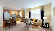 This single-floor residence is thoughtfully designed with an open-concept layout that seamlessly connects the living, dining, and kitchen areas. Warm yellow accent walls and neutral carpet throughout create a cohesive, welcoming ambiance, complemented by an abundance of natural light streaming through large windows dressed with bold navy and patterned drapery.

The kitchen is a standout feature, boasting warm wood cabinetry, dark granite countertops, stainless steel appliances, and a curved breakfast bar with pendant lighting overhead. In-unit laundry is also conveniently located within the kitchen area. The open dining area accommodates a round glass-top table and is illuminated by stylish track lighting. The adjacent living room offers generous space with large windows and wall-mounted television access.

The primary bedroom is spacious and bright, featuring multiple windows, plush carpeting, and a well-appointed en-suite bathroom with a glass-enclosed shower and tile surround. A second bedroom provides equal comfort with ample natural light and generous proportions. The second bathroom features a single vanity with wood cabinetry, a large mirror, and light tile flooring, maintaining the consistent, refined aesthetic found throughout the residence.