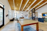 Located at 235 Verbena St in Charlotte, NC, this single-floor amenity space delivers a thoughtfully designed environment built for socializing, recreation, and relaxation. The interior is immediately distinguished by its soaring vaulted ceilings clad in warm wood planking, supported by exposed white painted beams and complemented by recessed lighting — creating a bright, airy atmosphere throughout.

Large windows along the perimeter flood the space with abundant natural light and offer views to the surrounding outdoor terrace, accessible via full-height glass doors. The main open floor plan is anchored by a central ping-pong table set atop a textured area rug over stone-tiled flooring, providing a focal point for recreational activity.

A comfortable lounge area features contemporary seating arrangements with leather sofas and accent chairs, framed by an open wood shelving unit that serves as a stylish room divider. A dramatic brick column adds industrial character, while curated wall art contributes to the space's modern aesthetic.

The kitchenette area is a standout feature, showcasing bold forest-green cabinetry paired with a striking blue marbled stone island with bar seating, white tile backsplash, and integrated appliances — ideal for casual dining and entertaining. A wall-mounted display screen adds a media element to this zone, enhancing the overall functionality of this well-appointed communal space.