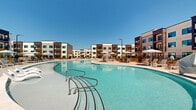 Located at 7135 Mount Zion Cir in Morrow, Georgia, this outdoor amenity space serves as the centerpiece of a contemporary residential community. The focal point is a generously sized freeform pool with a distinctive figure-eight shape, featuring crystal-clear turquoise water and a smooth concrete surround that provides ample space for lounging and recreation. The expansive pool deck is outfitted with numerous chaise lounge chairs and shaded seating areas beneath large market umbrellas, offering both sun-soaked and sheltered options for relaxation. A pergola structure at one end of the deck provides an additional gathering spot with covered seating. The entire pool area is enclosed by a sleek black wrought-iron fence with a gated entry, ensuring both safety and privacy. Surrounding the pool, young trees and manicured landscaping add a touch of greenery against the polished hardscape. The community's multi-story residential buildings frame the space on multiple sides, showcasing a modern architectural design with neutral facades, bold dark accent panels, large windows, and private balconies. A separate single-story clubhouse or leasing building with contemporary finishes and large glass storefronts is also visible on the grounds, further enhancing the community's well-appointed amenity offerings under the open Georgia sky.