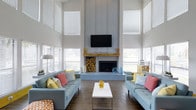 This single-story home in Irving offers a thoughtfully designed interior that balances modern aesthetics with a warm, inviting atmosphere. The standout living area immediately captures attention with its dramatically high ceilings that stretch upward, flanked by an abundance of large windows dressed with white blinds that flood the space with natural light. At the heart of the room sits a striking floor-to-ceiling fireplace clad in corrugated metal paneling, topped with a rustic wood mantel and complemented by a neatly stacked firewood display — a design feature that masterfully blends industrial and farmhouse elements. A wall-mounted flat-screen television is positioned above the mantel, adding a contemporary touch. Two plush, powder-blue sofas face each other across a sleek white coffee table, accented with vibrant coral, yellow, and gray throw pillows that inject a lively pop of color throughout the room. Dark hardwood flooring grounds the space and provides a rich contrast to the light-toned walls and furnishings. The overall ambiance is open, airy, and cheerful — a welcoming environment that combines striking architectural details with a carefully curated, colorful interior design palette.