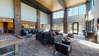 Located at 2100 Progress Pkwy in Schaumburg, Illinois, this single-floor amenity lounge is a thoughtfully designed communal space that blends industrial character with contemporary comfort. The architecture makes a striking impression through soaring double-height ceilings clad in warm wood paneling, dramatic exposed brick columns, and expansive floor-to-ceiling windows that flood the interior with abundant natural light and offer views of the surrounding outdoor areas.

The main lounge area is generously proportioned, featuring plush sectional sofas, accent chairs, and round coffee tables arranged to encourage both socializing and relaxation. A large wall-mounted television and fireplace anchor one end of the lounge, while a billiard table adds a recreational focal point nearby. Pendant lighting throughout creates a warm, inviting ambiance during any time of day.

A fully equipped kitchen bar provides wood cabinetry, a dark stone backsplash, a large island with seating, a mounted television, and stainless steel appliances — ideal for entertaining and casual gatherings. Adjacent areas feature banquette-style seating with pendant lighting, offering more intimate nooks within the open floor plan.

The space transitions seamlessly between zones via light hardwood flooring and patterned carpet, while sculptural artwork and curated decor add an elevated, gallery-like quality throughout the entire lounge.