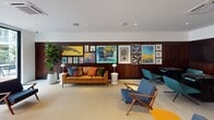 Located at 1055 W Town and Country Rd in Orange, California, this single-floor amenity lounge is a thoughtfully designed communal space that blends mid-century modern aesthetics with contemporary comfort. The open-plan layout is anchored by light-toned tile flooring and bright white ceilings fitted with recessed lighting and ceiling-cassette HVAC units, creating a clean and airy atmosphere throughout.

Seating areas are generously distributed across the space, featuring a rich cognac leather sofa accented with colorful throw pillows, solid wood-framed lounge chairs in navy and slate blue upholstery, and sculptural round ottoman poufs that add an organic touch. A curated gallery wall of bold, vibrant artwork lines the dark wood-paneled accent wall, adding significant visual character to the room.

A dedicated game zone houses a substantial shuffleboard table crafted from natural wood, positioned against a dark paneled wall adorned with a series of abstract floral paintings. Adjacent to the game area, a built-in media and display wall features teal-painted lower cabinetry, warm-lit open shelving filled with books and decorative objects, and a mounted flat-screen television.

Large floor-to-ceiling sliding glass doors open directly to an outdoor terrace, flooding the interior with natural light and providing seamless indoor-outdoor connectivity. Lush indoor greenery, including a prominent fiddle-leaf fig tree, enhances the inviting and sophisticated ambiance of the space.