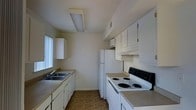 This two-floor space includes a galley-style kitchen designed for efficient use of space. The kitchen is equipped with white cabinetry lining both sides of the room, providing ample storage above and below the countertops. A double-basin sink is positioned along one wall beneath a window dressed with vertical blinds, allowing natural light to filter in. On the opposite wall, a white electric coil-top range and oven sit alongside a full-size white refrigerator, complemented by an overhead range hood for ventilation. A flush-mounted ceiling light fixture provides consistent overhead illumination throughout the kitchen. The flooring features a parquet-style wood pattern, adding a classic touch to the space. Upper cabinets extend toward the ceiling, maximizing vertical storage capacity. The neutral wall tones and white cabinetry create a clean, understated aesthetic throughout the kitchen. The overall layout is straightforward and practical, with clearly defined cooking, storage, and prep zones. As a two-floor property, additional living spaces are distributed across both levels, offering a functional and organized living arrangement.