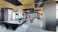 Nouveau Lounge, located at 969 Commerce St in downtown Fort Worth, Texas, is a sophisticated and generously proportioned entertainment space designed with a bold, contemporary aesthetic. The open-plan layout is anchored by a striking geometric-patterned tile floor that flows throughout the main lounge area, complemented by a dramatic ceiling design featuring gold latticed panels set against an exposed black industrial framework, with pendant lighting adding layers of ambiance.

The main lounge area is thoughtfully arranged with multiple seating vignettes, including plush teal sofas, lounge chairs, and daybeds, all oriented toward large floor-to-ceiling windows that frame panoramic views of the Fort Worth skyline. A full-size billiards table serves as a focal point in the entertainment zone, accompanied by a warm wood-paneled accent wall and a mounted flat-screen display. A sculptural marble column with an integrated aquarium feature adds a dramatic visual centerpiece to the space.

The dining area showcases a dark granite table surrounded by rich cognac leather chairs beneath an elegant cluster of pendant lights. The fully equipped kitchen features dark wood cabinetry, granite countertops with brass-accented details, and high-end appliances. Throughout the space, curated artwork and refined furnishings reinforce the lounge's upscale, modern character.